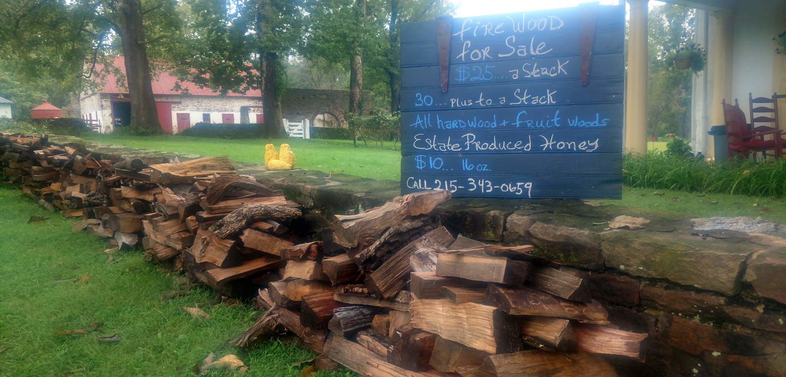 sign selling firewood with stacks of wood against stone wall with barn in distant background