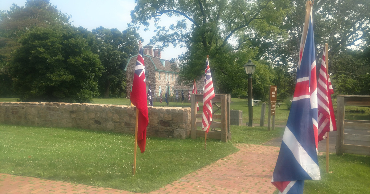 colonial flags in foreground with stone wall behind and stone house further back in center