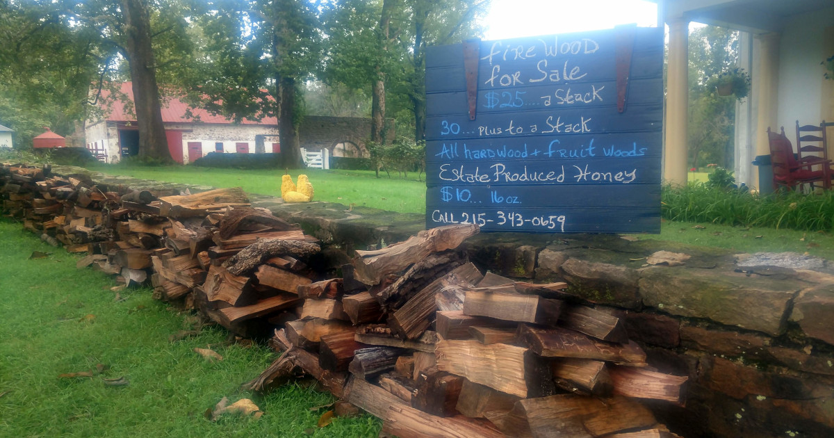 sign selling firewood with stacks of wood against stone wall with barn in distant background